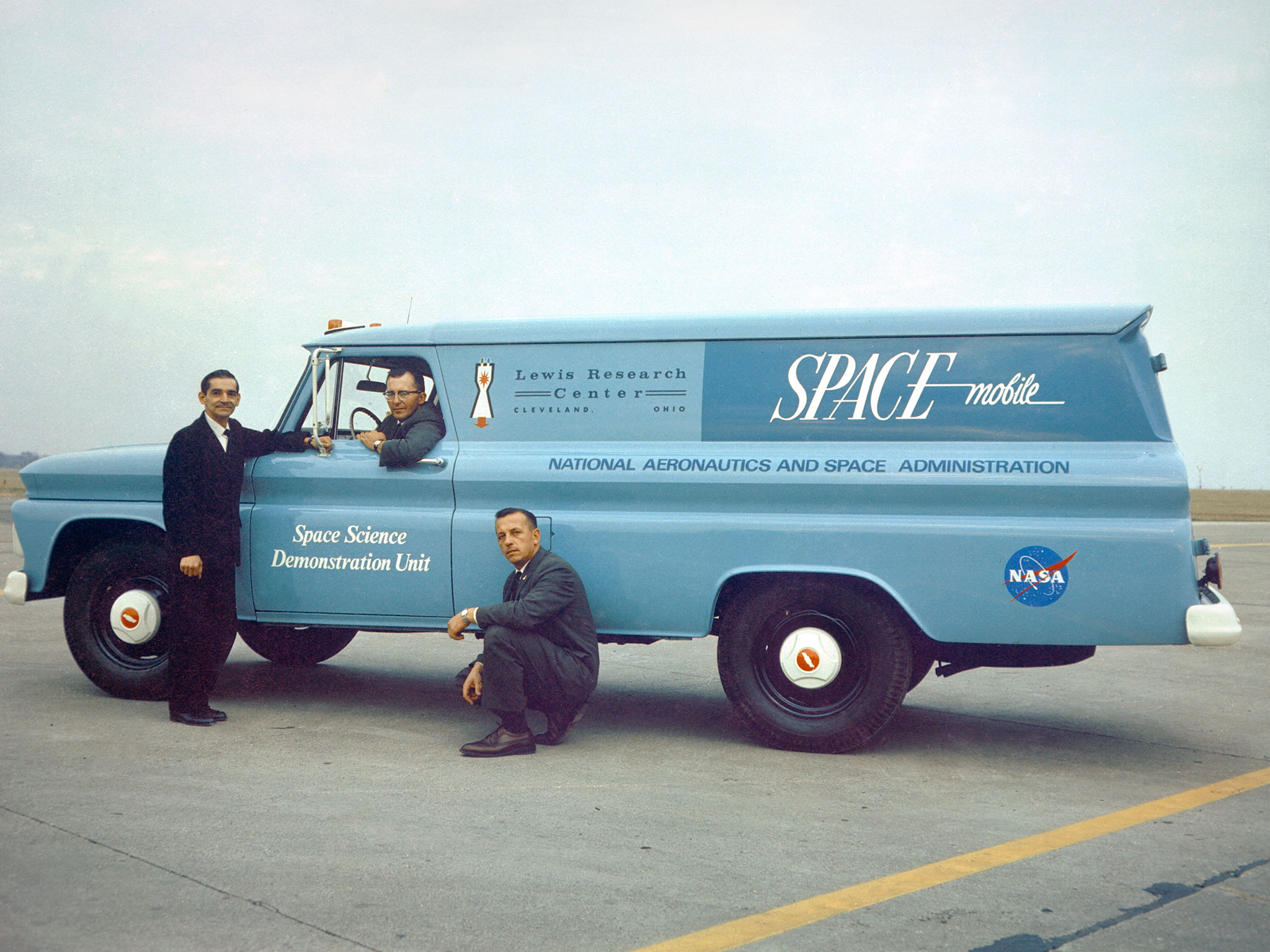 One man sits in the driver seat of a light blue van, while two other men stand and crouch near him on the outside. They are all wearing dark suits. The van door has white writing on it that reads "Space Science Demonstration Unit." The van also has writing and several logos, including the NASA meatball, the Lewis Research Center name and logo, as well as the "Spacemobile" logo ("space" is written in all caps), and finally, "National Aeronautics and Space Administration."